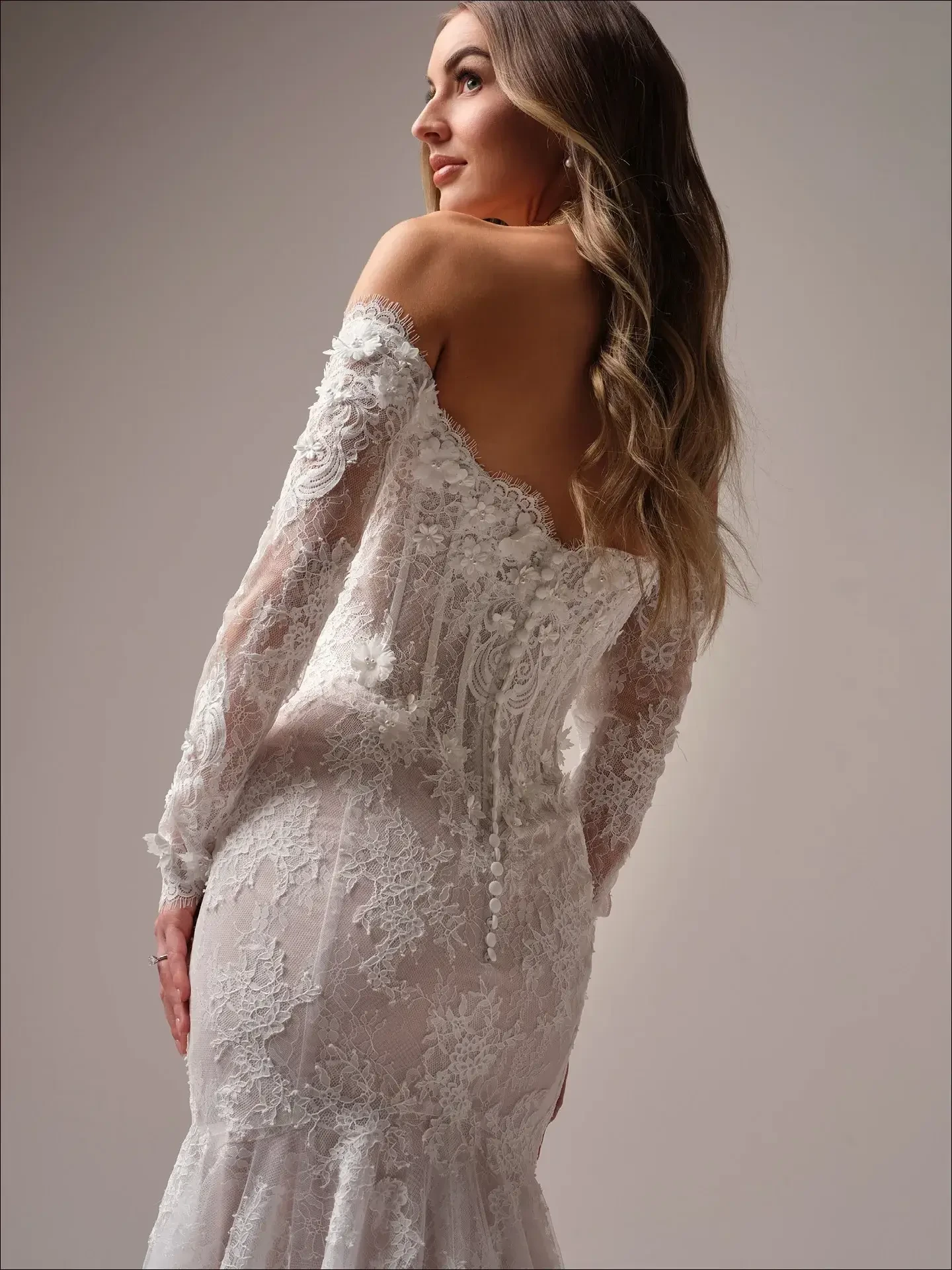 Woman in a lace off-shoulder wedding dress gazes over her shoulder. The dress features intricate lace patterns and buttons, conveying elegance and romance.