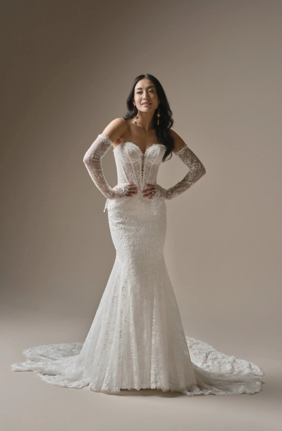 A woman in a white wedding dress with lace details and long, sheer sleeves, standing confidently against a neutral background.