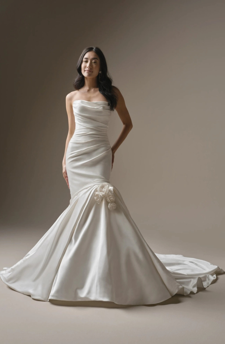 A woman wearing a strapless white wedding gown with a mermaid silhouette, featuring delicate floral embellishments at the waist. The background is a neutral tone to highlight the dress.