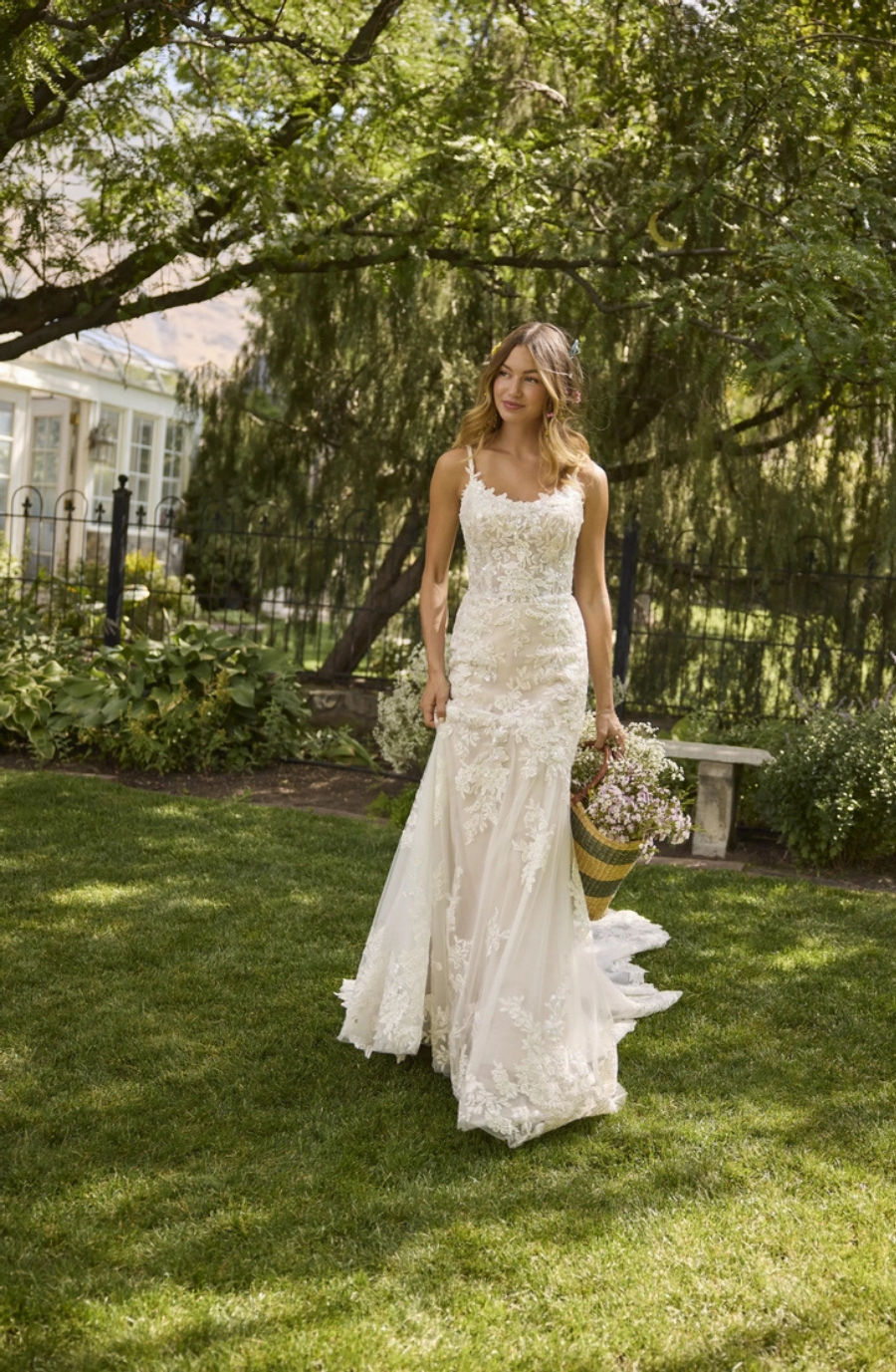 A woman wearing an elegant wedding dress, standing outdoors in a garden. She is holding a bouquet of flowers and has long hair cascading down her shoulders. The background features greenery and trees.