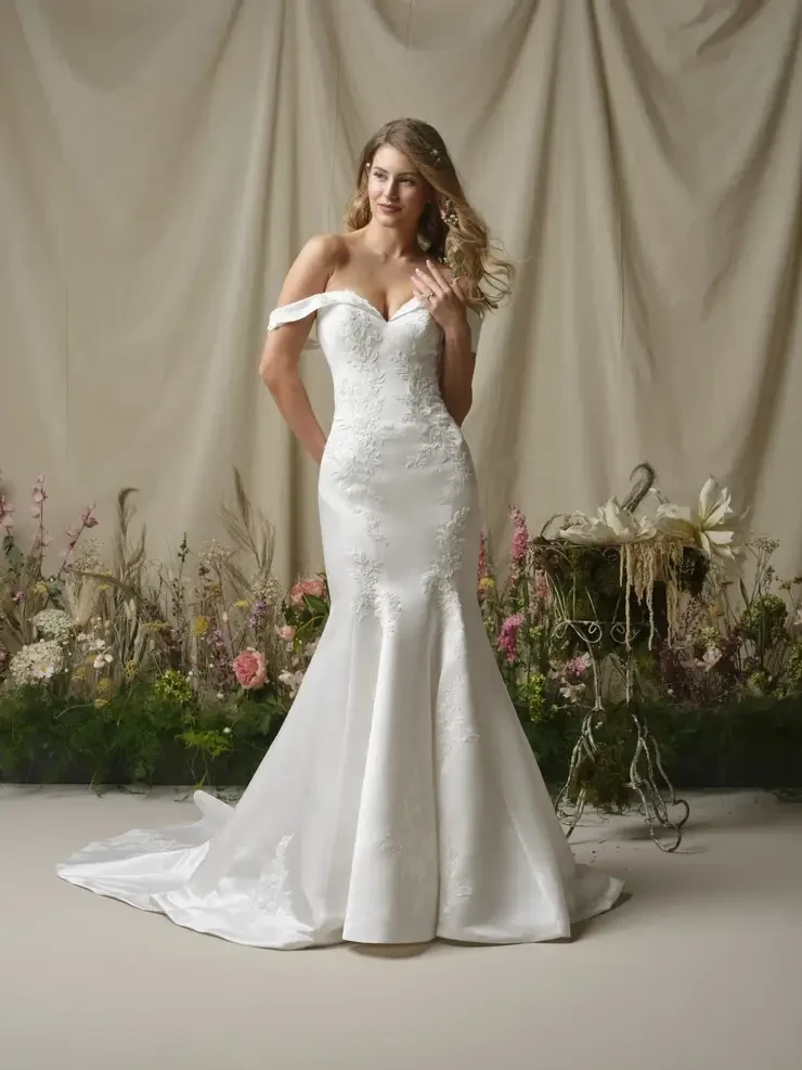 A woman wearing an elegant off-the-shoulder wedding dress stands in a floral setting with a neutral background.