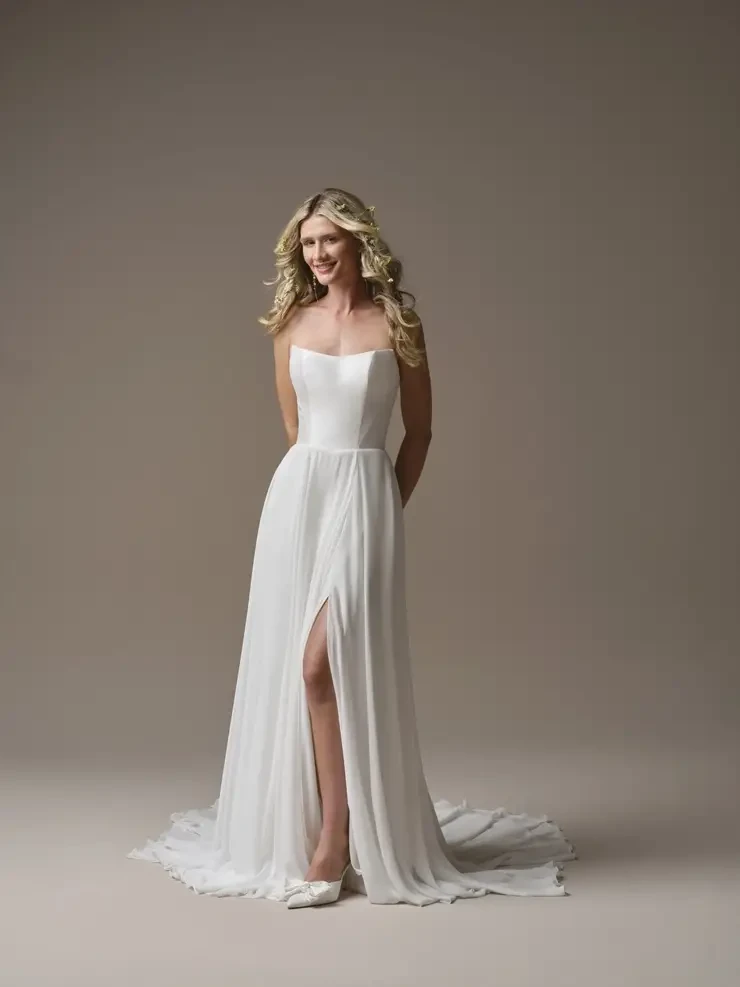 A woman wearing a simple, elegant white gown with a high slit, standing against a neutral background. Her long hair is styled in loose waves.