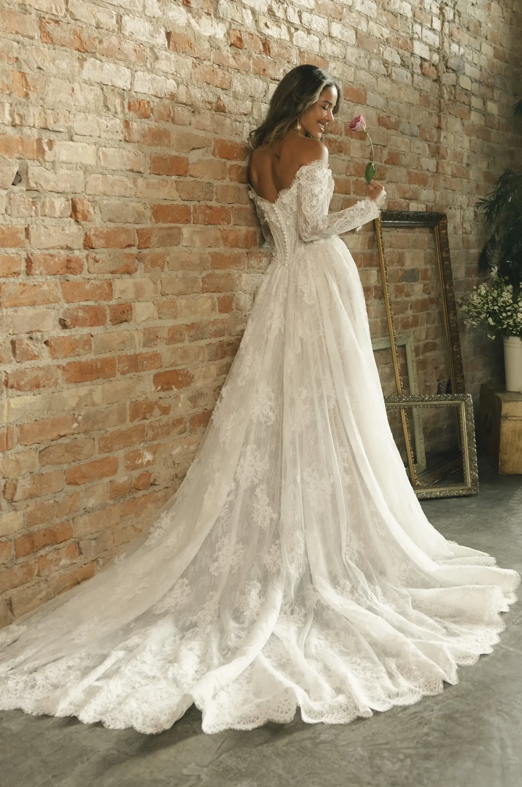 A woman in an elegant, lace wedding gown stands against a rustic brick wall, holding a pink rose, with empty frames and soft lighting conveying romance.