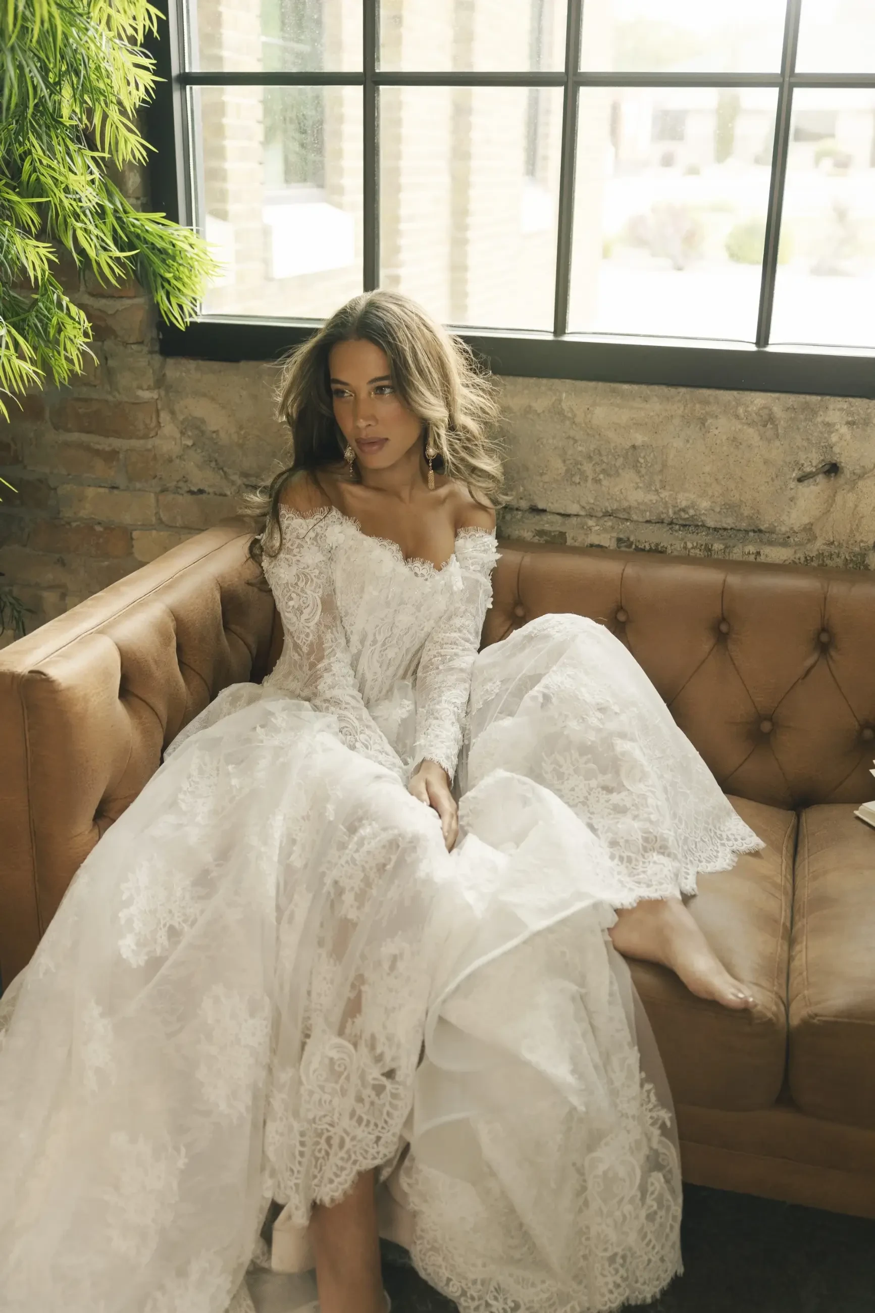 Bride in a lacy, off-shoulder wedding dress sits on a tan leather couch, gazing thoughtfully out a window. Rustic brick walls and green plant nearby.