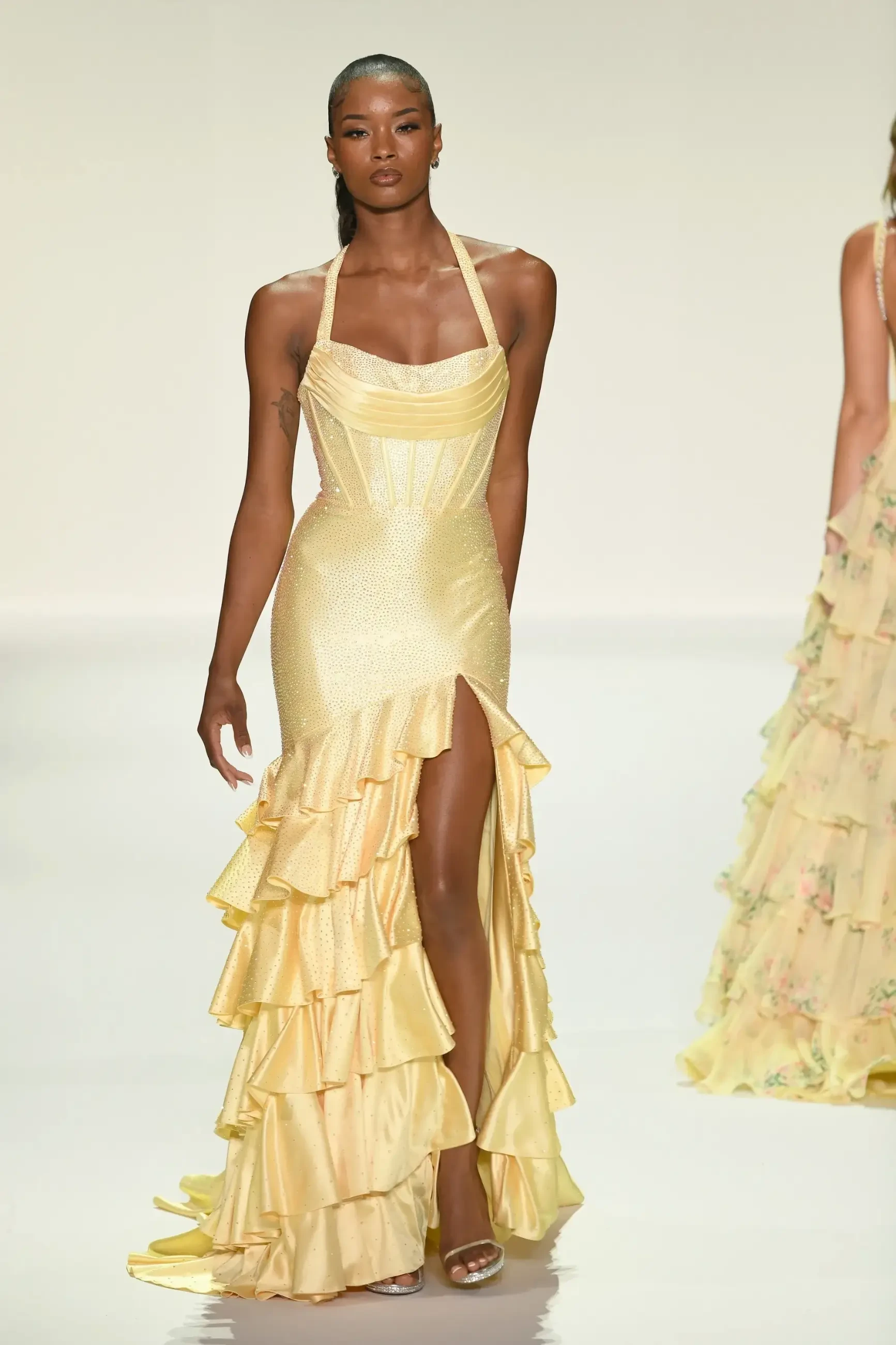 A model walks down the runway wearing a yellow evening gown with ruffles and a thigh-high slit.