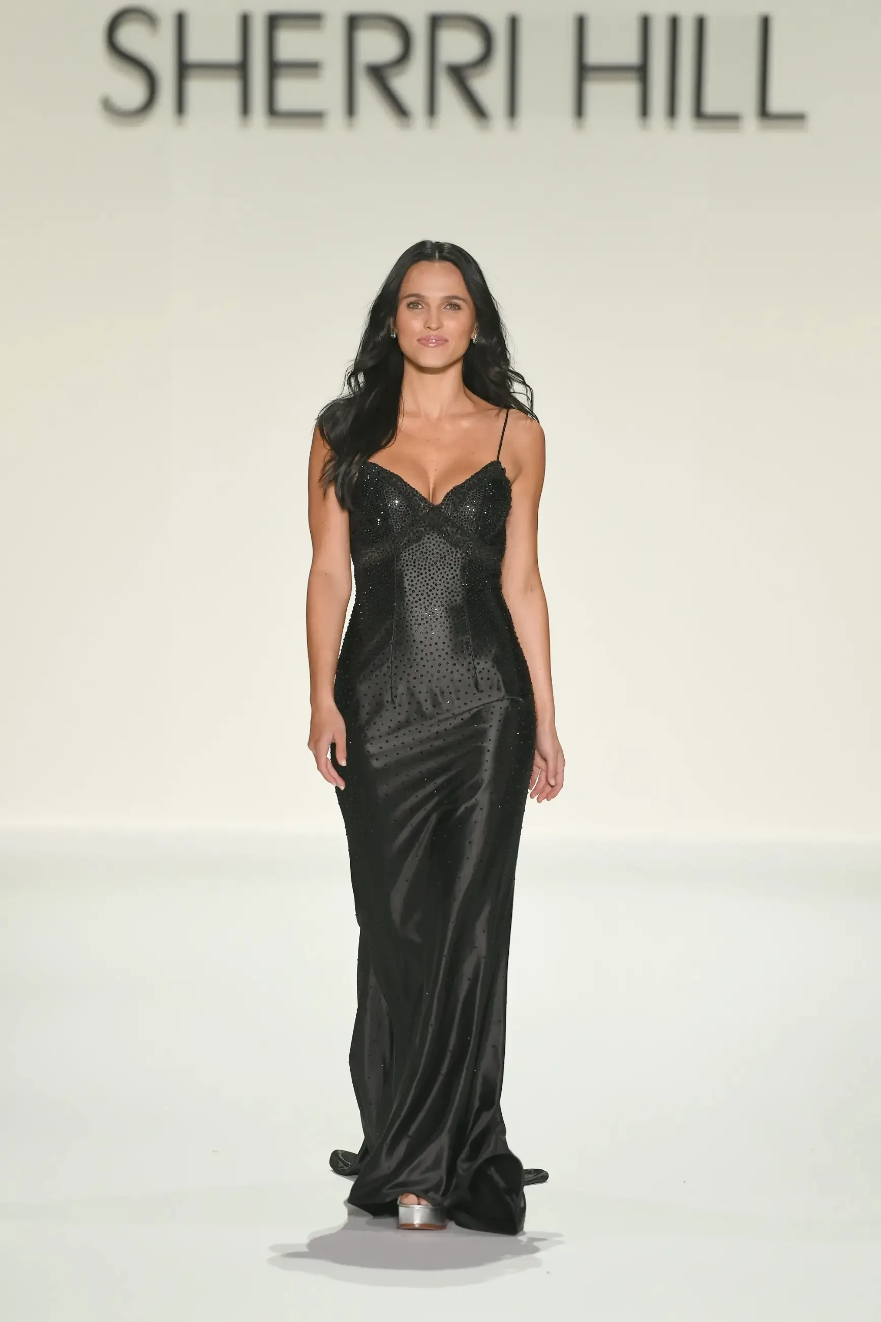 A model in a shimmering black gown walks confidently down the runway, exuding elegance, with 
