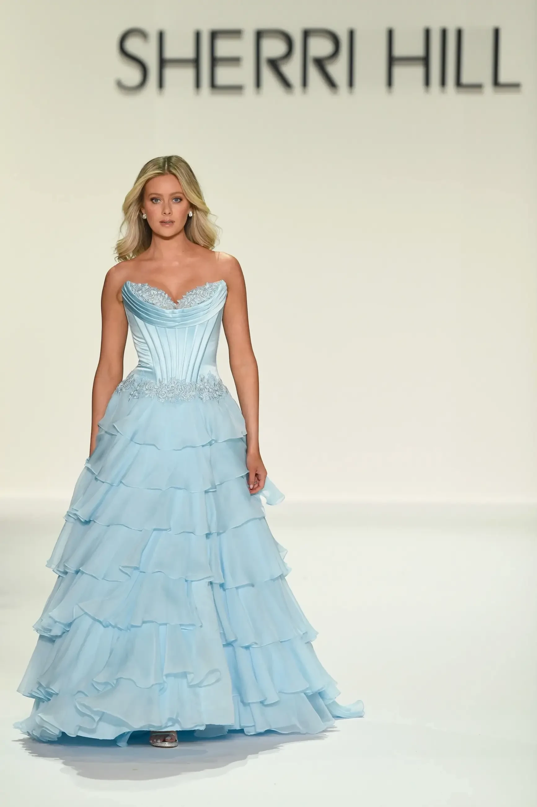 A model walking down the runway in a light blue gown with ruffles, featuring a sweetheart neckline, during a Sherri Hill fashion show.