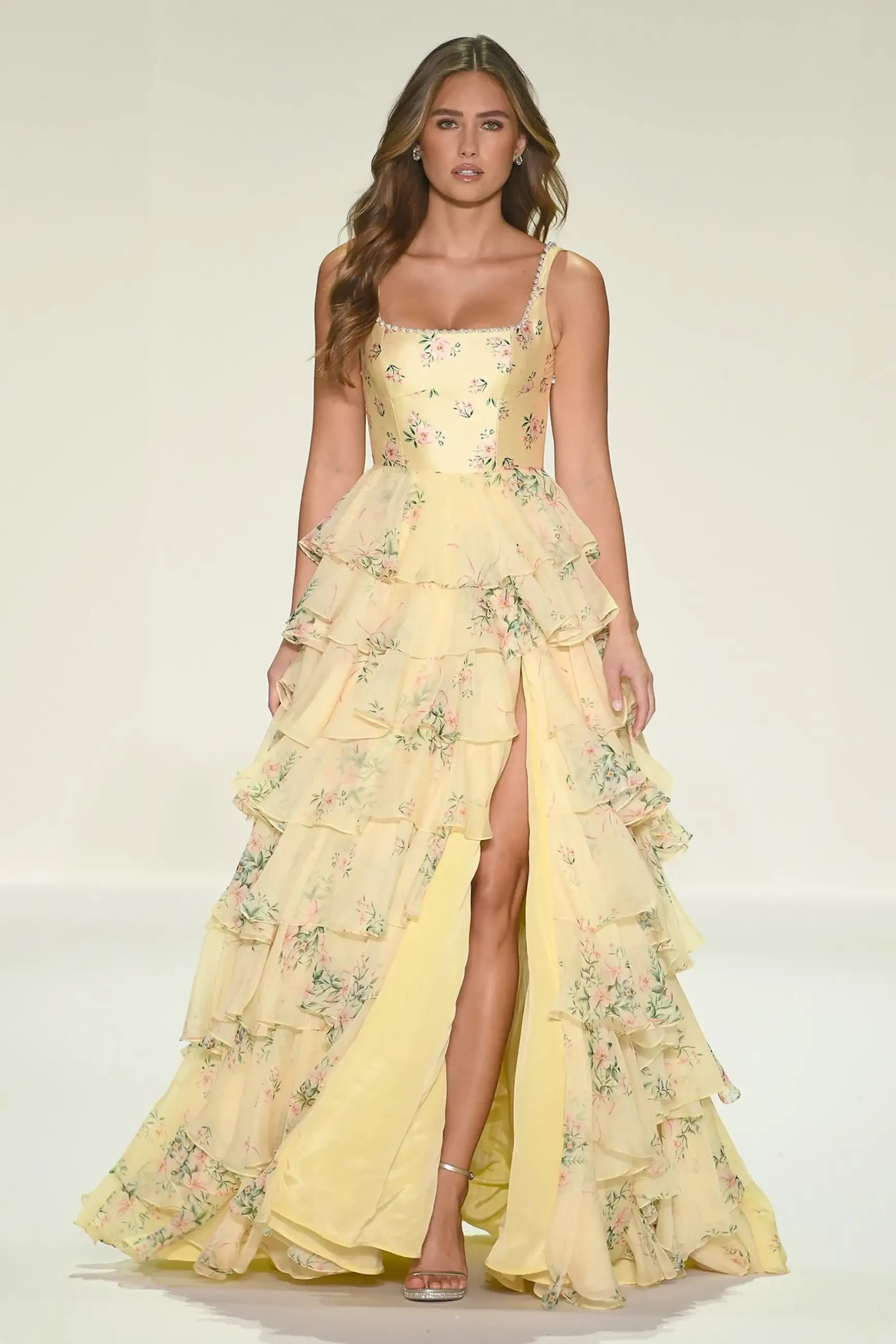 A model wearing a yellow floral gown with multiple ruffles and a thigh-high slit, walking down the runway.
