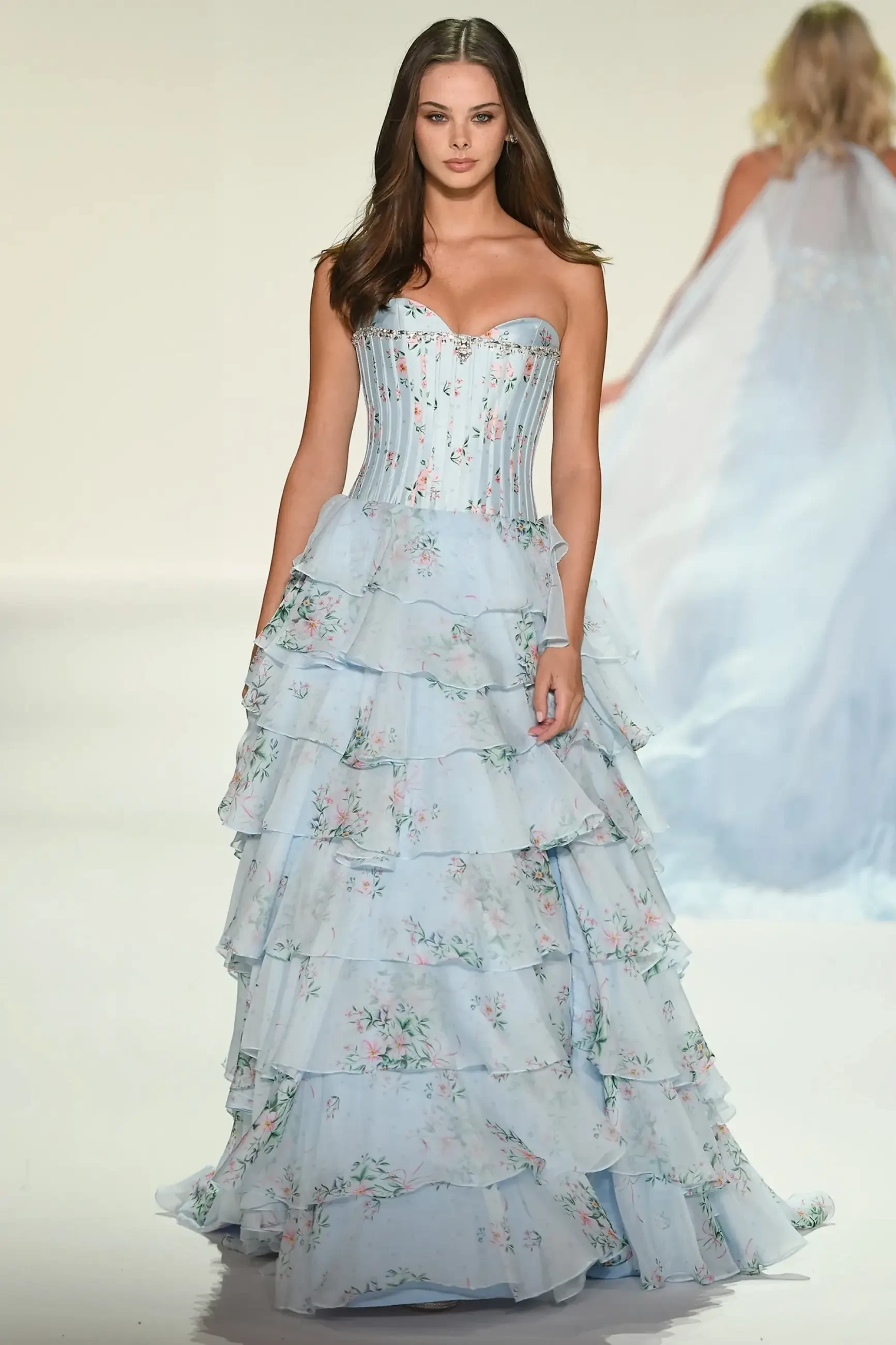 A model walks the runway wearing a strapless, light blue gown with floral patterns and layered, ruffled skirts.