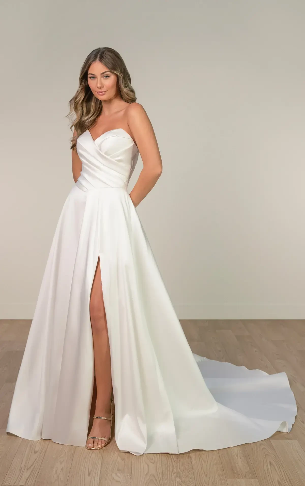 A woman in a strapless white satin wedding gown with a sweetheart neckline and high slit stands on a wooden floor, exuding elegance and confidence.