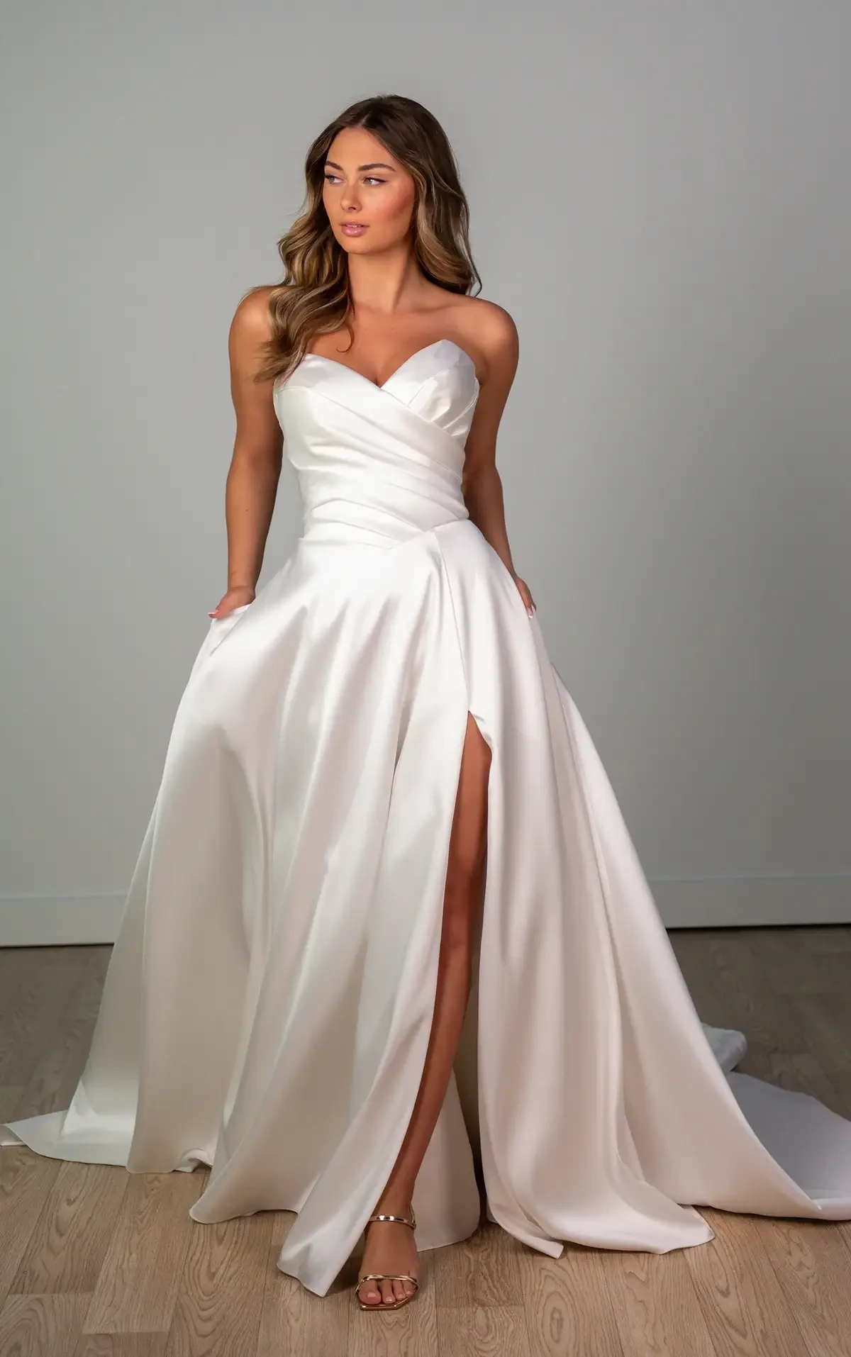A woman in a strapless white satin wedding gown with a sweetheart neckline and high slit stands on a wooden floor, exuding elegance and confidence.