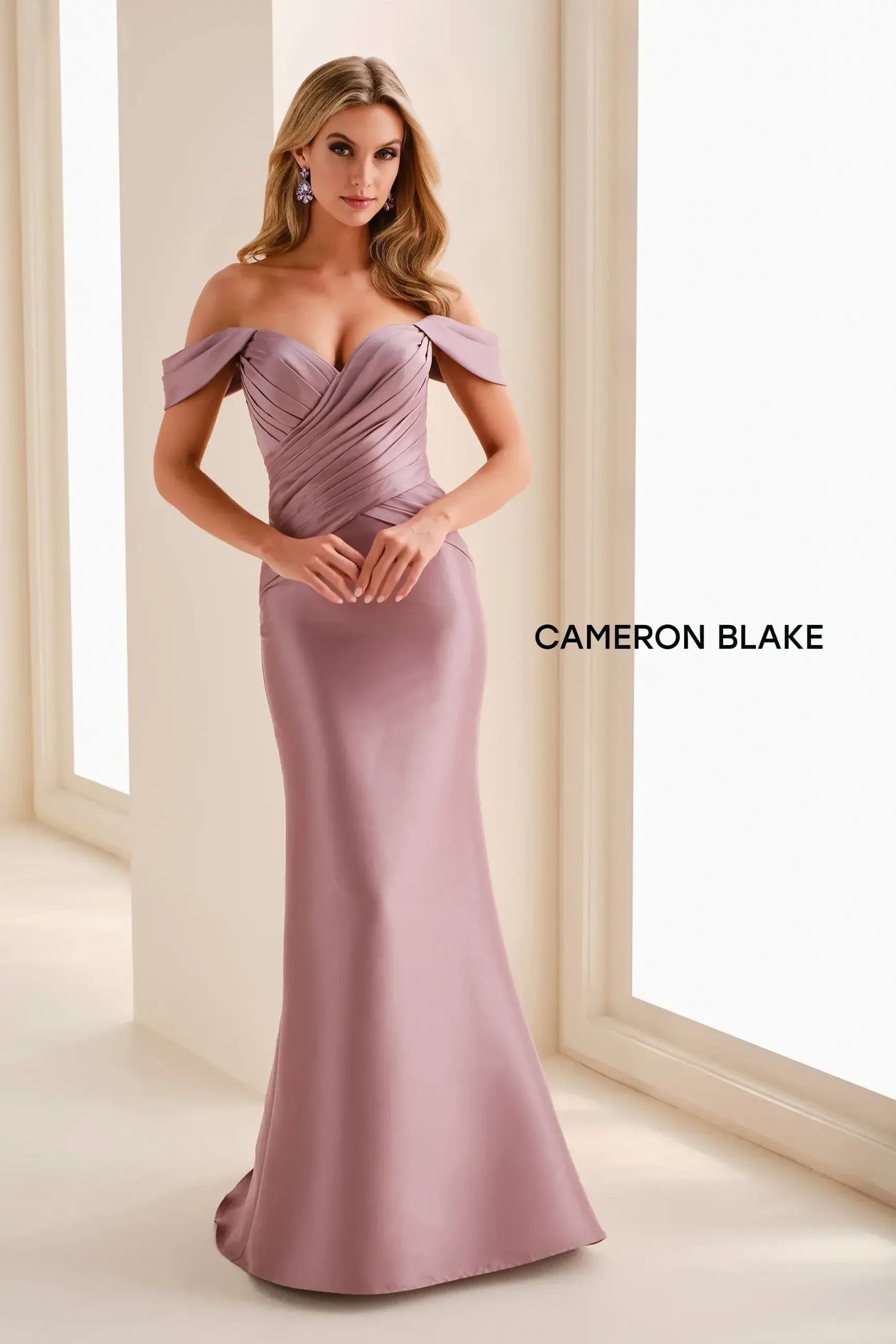 A woman in an elegant, fitted evening gown with an off-the-shoulder design, standing in a well-lit room. The gown is a soft lavender color and she is posing with her hands clasped together.