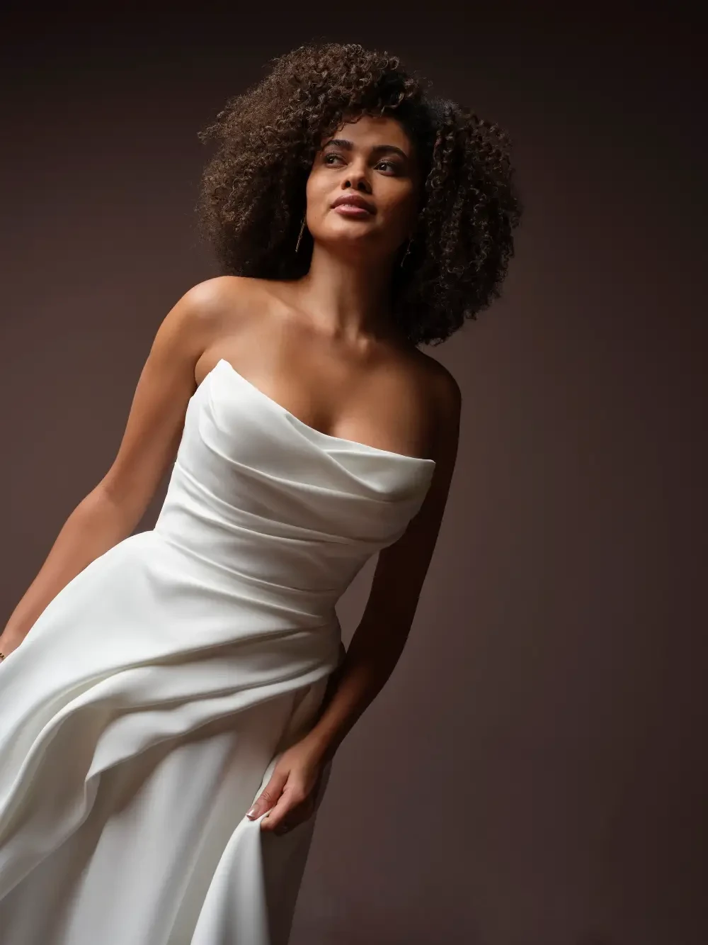 A woman in a strapless white dress, with curly hair, posing confidently against a dark background.