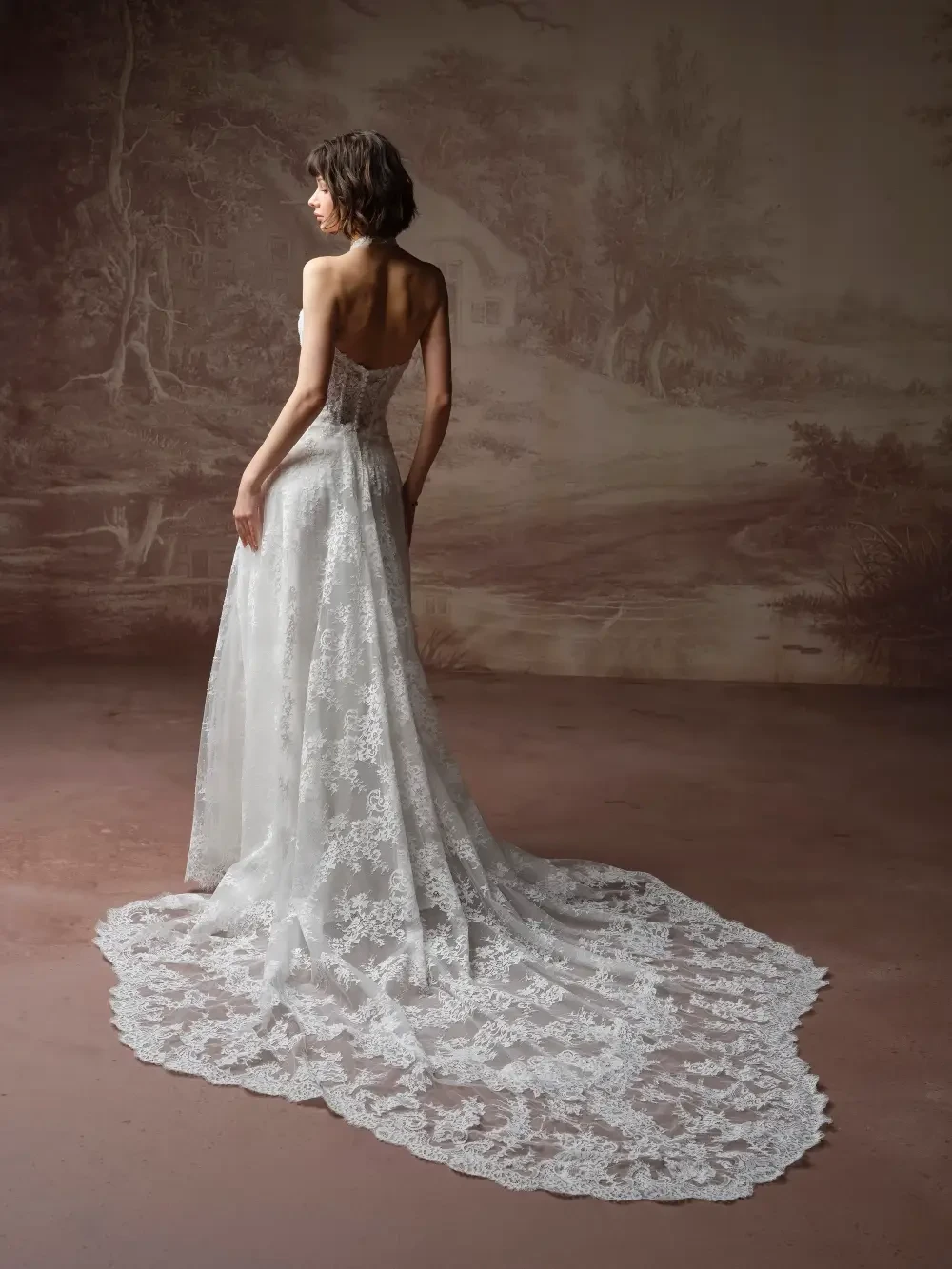 A model wearing a long, elegant lace wedding dress, standing with her back to the camera in a softly lit, artistic setting.