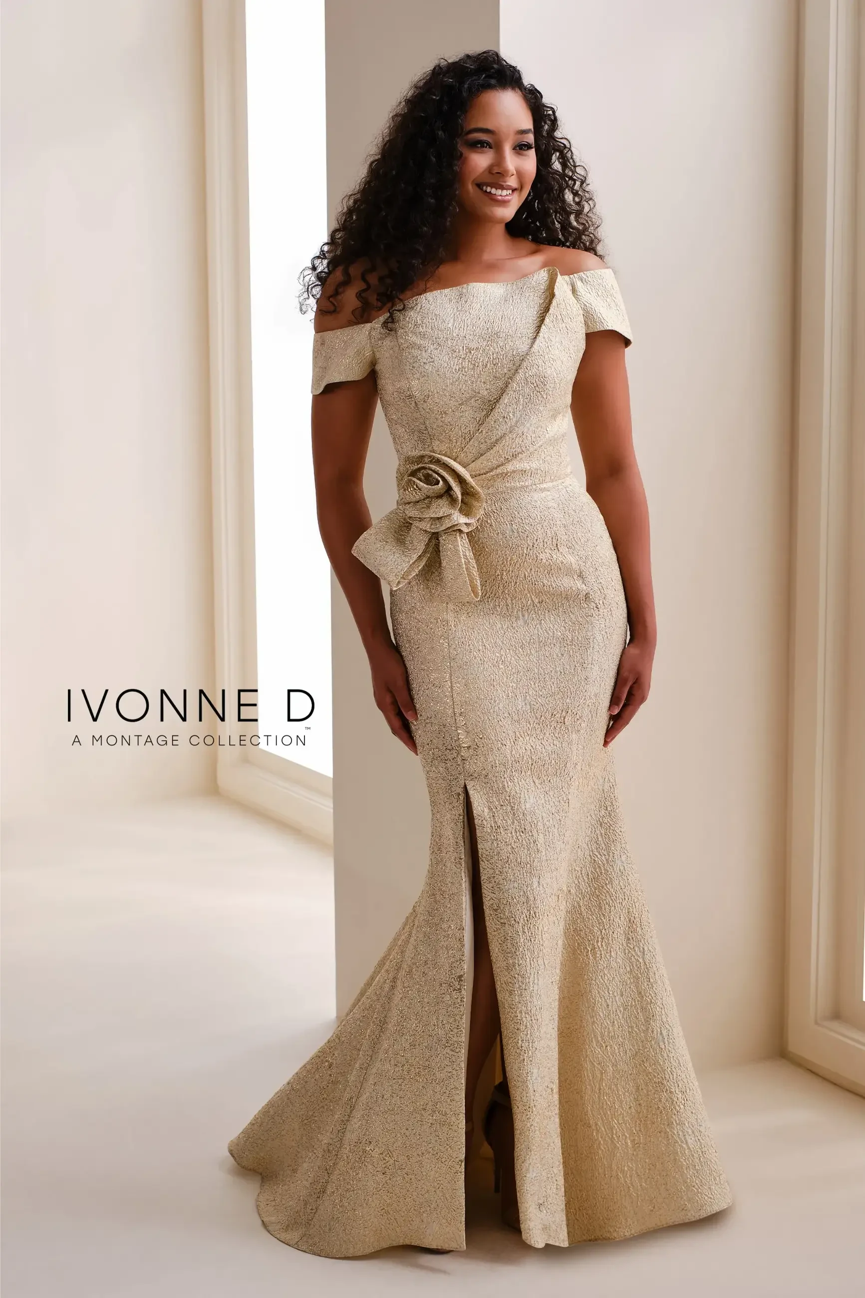 A woman wearing an elegant off-the-shoulder dress with a fitted silhouette, featuring a decorative bow at the waist and a side slit. She has long, curly hair and is standing against a light-colored background.