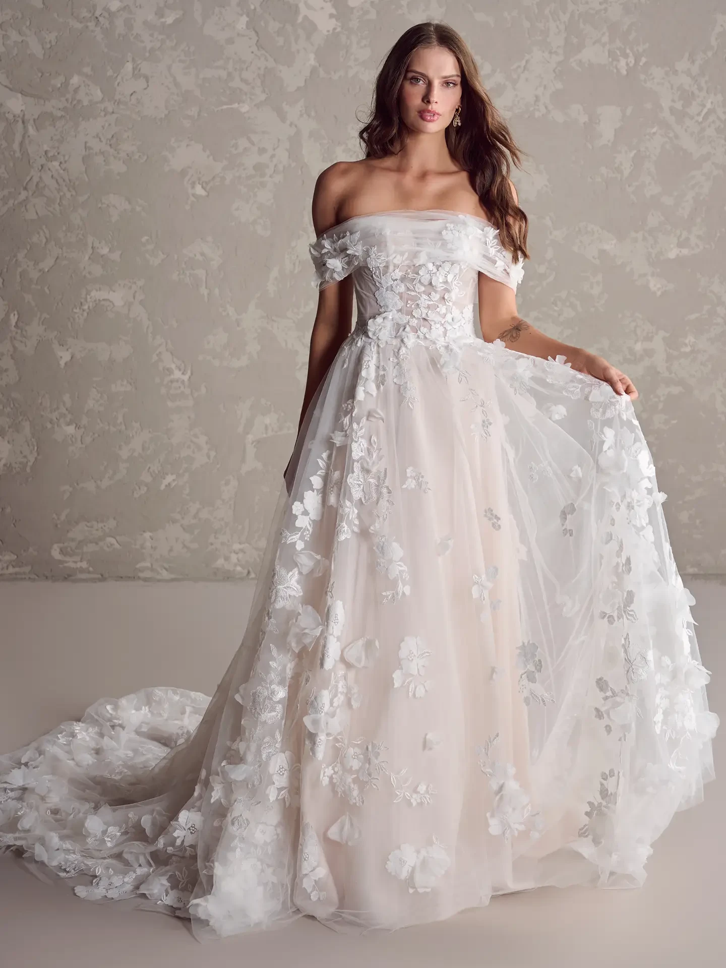 Brunette woman in an off-shoulder, floral-embroidered wedding gown standing against a textured gray wall. The elegant dress flows gracefully.