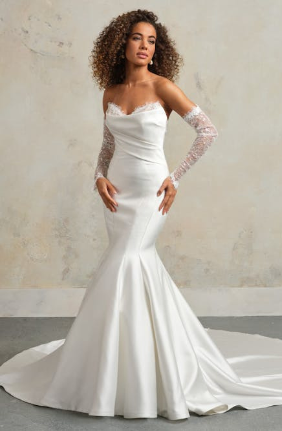 A woman in a strapless, form-fitting white wedding dress with lace gloves stands against a textured beige wall. The dress flares elegantly at the bottom.