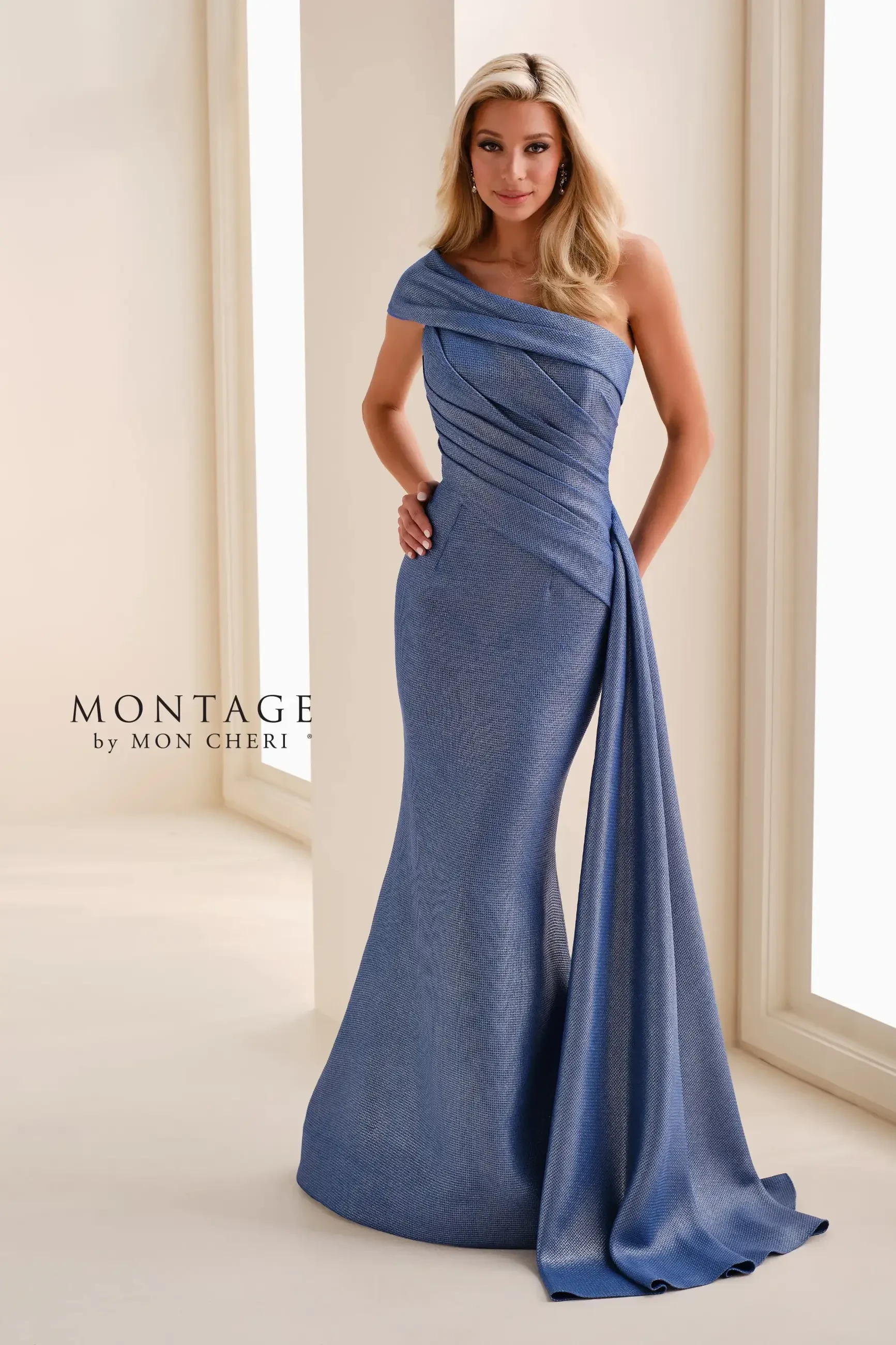 A woman wearing a one-shoulder, floor-length evening gown in blue, standing elegantly in a well-lit indoor setting. The dress features draped fabric design and a fitted silhouette.
